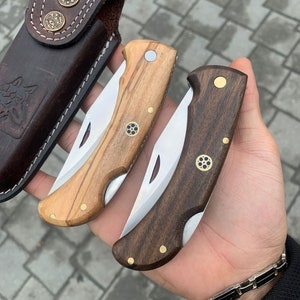 May include: Two folding knives with wooden handles, one light and one dark, and stainless steel blades. A brown leather sheath with decorative metal studs and a Celtic knot design is also visible. The knives are held in a hand.