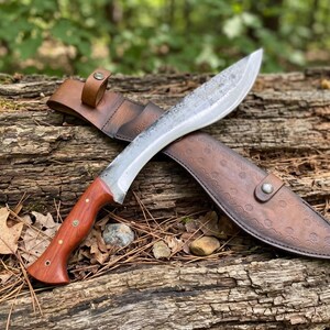 May include: A large, curved blade knife with a wooden handle and a leather sheath. The blade is silver and the handle is a reddish-brown color. The leather sheath is brown and has a decorative pattern. The knife is resting on a weathered log.