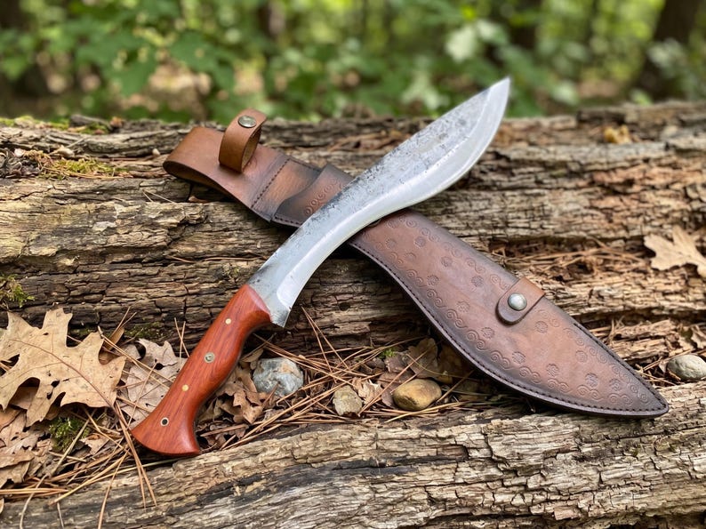 May include: A large, curved blade knife with a wooden handle and a leather sheath. The blade is silver and the handle is a reddish-brown color. The leather sheath is brown and has a decorative pattern. The knife is resting on a weathered log.