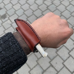 May include: A small, handcrafted knife with a bone handle and a brown leather sheath. The sheath is stitched with black thread and is worn on the wrist. The knife is a light color and has a small metal rivet.