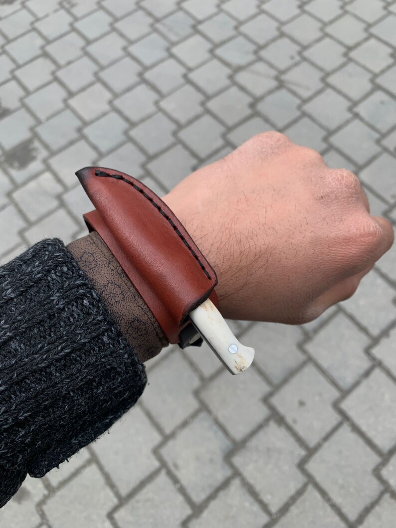 May include: A small, handcrafted knife with a bone handle and a brown leather sheath. The sheath is stitched with black thread and is worn on the wrist. The knife is a light color and has a small metal rivet.