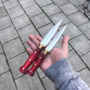 May include: Two small, silver, double-edged daggers with red wooden handles. The handles have white dots and gold accents.