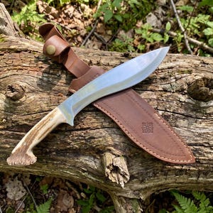 May include: A large, curved hunting knife with a polished steel blade and a handle made of antler material. The knife rests in a brown leather sheath with a decorative embossed design, secured by a leather strap and button.