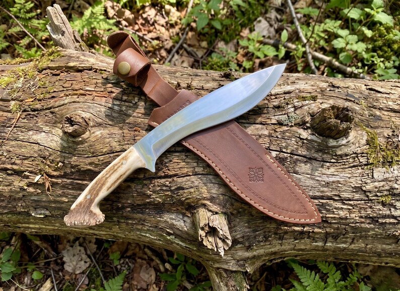 May include: A large, curved hunting knife with a polished steel blade and a handle made of antler material. The knife rests in a brown leather sheath with a decorative embossed design, secured by a leather strap and button.