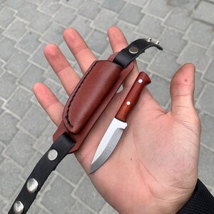 May include: A small, handcrafted knife with a wooden handle and a silver blade, accompanied by a brown leather sheath and a black leather strap with silver snaps. The knife is designed for outdoor use.