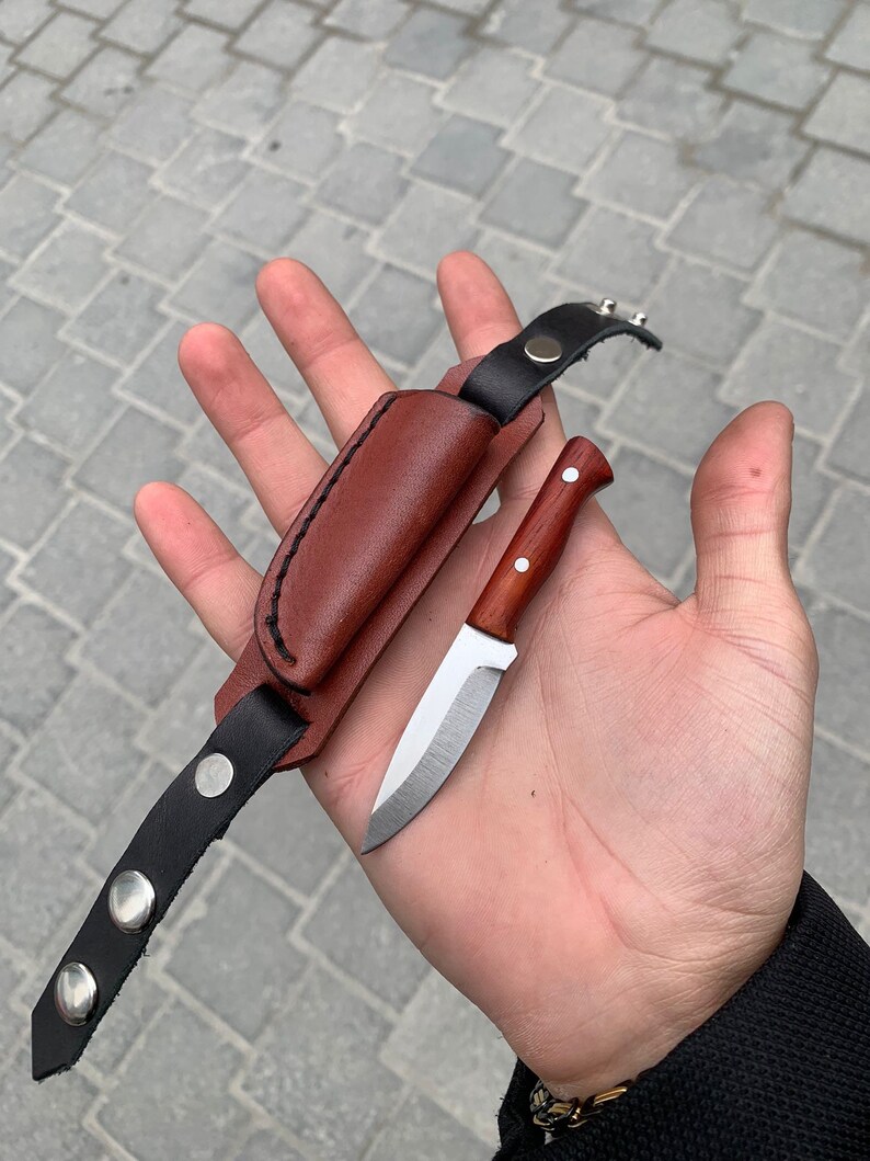 May include: A small, handcrafted knife with a wooden handle and a silver blade, accompanied by a brown leather sheath and a black leather strap with silver snaps. The knife is designed for outdoor use.