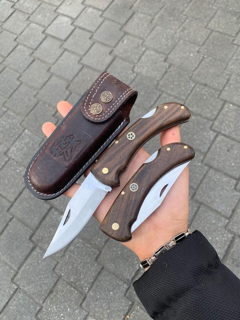 May include: Two folding knives with wooden handles and stainless steel blades, alongside a brown leather sheath. The knives have brass accents and a decorative emblem. The sheath features a wolf design and brass button closures.
