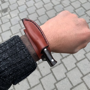 Può includere: Un piccolo coltello con manico scuro e fodero in pelle marrone. Il fodero è cucito con filo nero ed è attaccato a un cinturino da polso. Il coltello è mostrato sul polso di una persona.