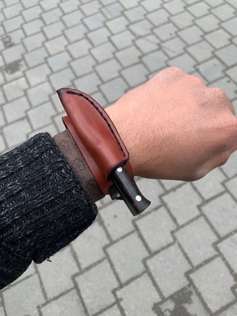 May include: A small, dark-handled knife with a brown leather sheath. The sheath is stitched with black thread and is attached to a wrist strap. The knife is shown on a person's wrist.
