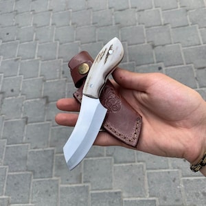 May include: A handmade hunting knife with a polished steel blade and a white handle with brown accents. The knife is paired with a brown leather sheath with decorative tooling.