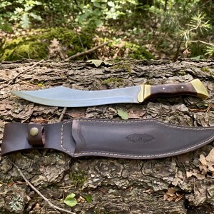 May include: A curved steel blade knife with a dark brown wooden handle and brass bolster, accompanied by a matching dark brown leather sheath featuring decorative embossed detailing, a snap button closure, and white contrast stitching.