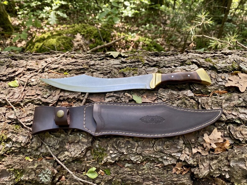 May include: A curved steel blade knife with a dark brown wooden handle and brass bolster, accompanied by a matching dark brown leather sheath featuring decorative embossed detailing, a snap button closure, and white contrast stitching.