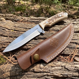 May include: A hunting knife with a silver blade and a handle made of bone-like material. The knife rests next to a brown leather sheath with a floral design and a button clasp. The knife and sheath are on a weathered log.