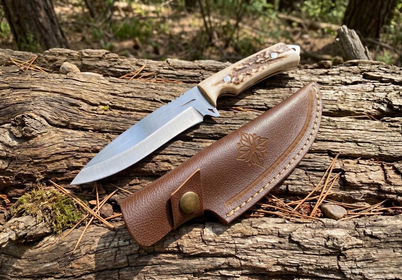 May include: A hunting knife with a silver blade and a handle made of bone-like material. The knife rests next to a brown leather sheath with a floral design and a button clasp. The knife and sheath are on a weathered log.