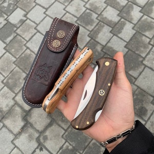 May include: Two folding pocket knives with wooden handles and a leather sheath. One knife has a light wood handle and the other has a dark wood handle. The sheath is brown with decorative metal buttons and a wolf emblem.