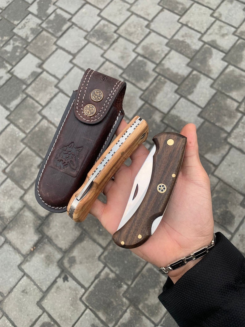 May include: Two folding pocket knives with wooden handles and a leather sheath. One knife has a light wood handle and the other has a dark wood handle. The sheath is brown with decorative metal buttons and a wolf emblem.
