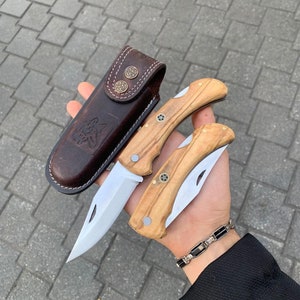 May include: Two folding knives with wooden handles and stainless steel blades, along with a brown leather sheath. The sheath features a wolf design and decorative metal studs. The knives are held in a hand.