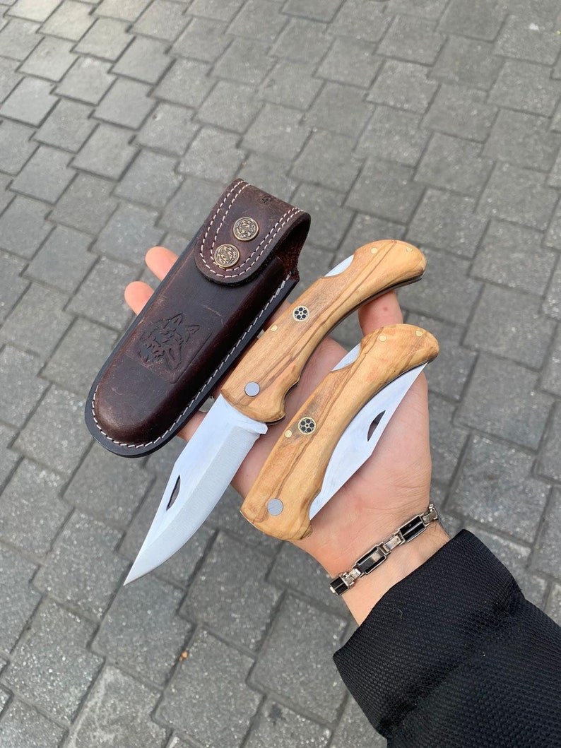 May include: Two folding knives with wooden handles and stainless steel blades, along with a brown leather sheath. The sheath features a wolf design and decorative metal studs. The knives are held in a hand.