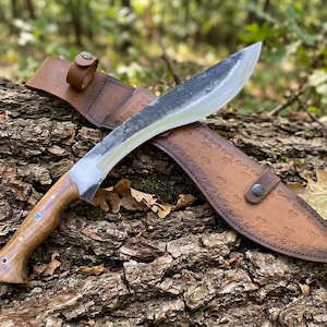 May include: A large, curved blade knife with a wooden handle and a leather sheath. The blade has a silver finish, and the handle is a light brown color. The sheath is brown and has a decorative pattern. The knife is resting on a log.