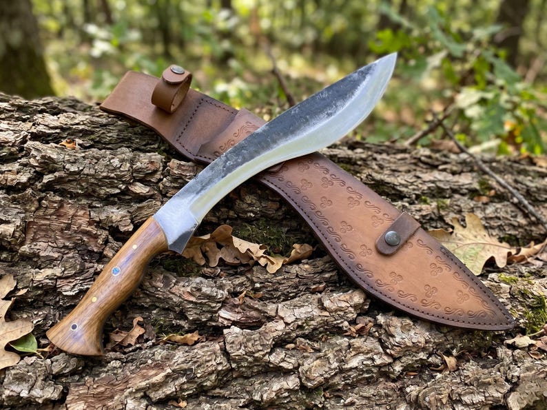 May include: A large, curved blade knife with a wooden handle and a leather sheath. The blade has a silver finish, and the handle is a light brown color. The sheath is brown and has a decorative pattern. The knife is resting on a log.
