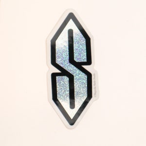 May include: A silver holographic sticker with a black outline of the letter "S" in a diamond shape. The sticker has a glittery finish and the text "Magnoila Mark's Studio" is printed on the right side.