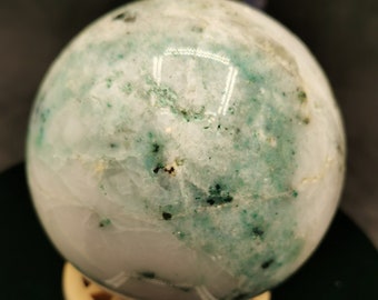 May include: A polished, spherical gemstone with a mix of white and green hues. The orb is speckled with darker green inclusions and rests on a cream-colored, geometric stand. The overall effect is a natural, decorative item.