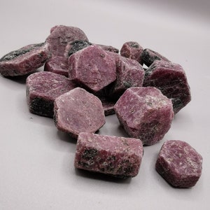 May include: A pile of rough, pink ruby gemstones. The rubies are various sizes and shapes, with some having a hexagonal shape.