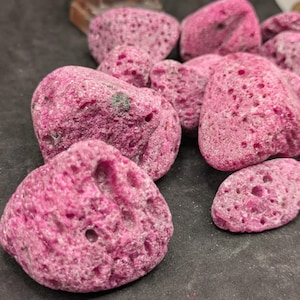 May include: A cluster of porous, irregularly shaped, magenta-pink rocks. The rocks have a rough texture with numerous small holes and varying sizes. The background is a dark, neutral colour, providing contrast to the vibrant pink stones.