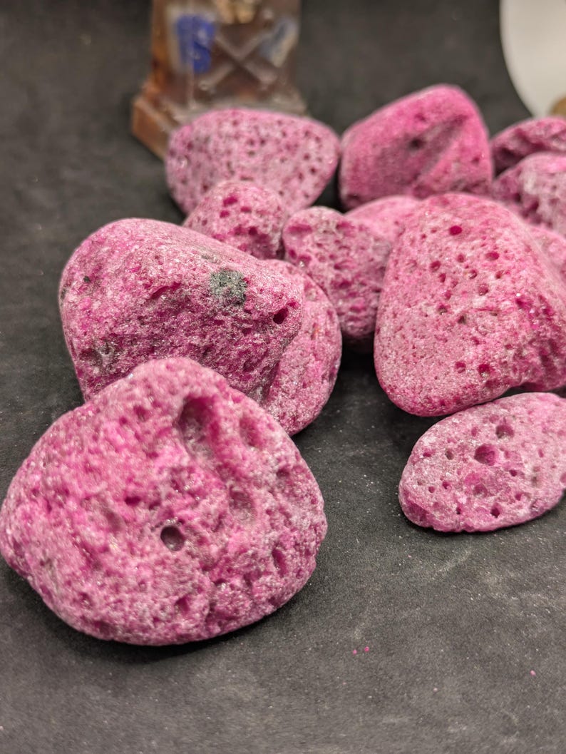 May include: A collection of porous, irregularly shaped, hot pink rocks. The rocks have a rough texture with numerous small holes and varying sizes. The background is a dark, neutral color, providing contrast to the vibrant pink stones.