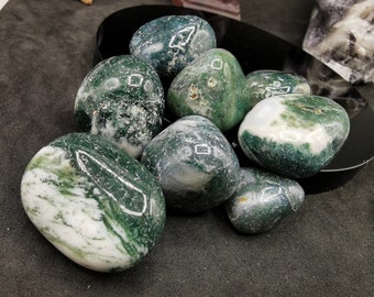 May include: A collection of polished moss agate stones. The stones are oval-shaped, with a mix of translucent white and deep green patterns. The stones are arranged on a black surface, with some stones reflecting light.