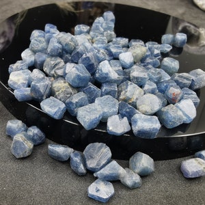 May include: A collection of rough, blue sapphire gemstones. The sapphires are various sizes and shapes, with a slightly cloudy appearance. The gemstones are displayed on a black surface.