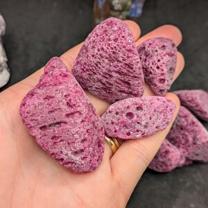 May include: A hand holds several pieces of pink, porous rock. The rocks vary in size and shape, with a textured surface and a deep pink color. The background is dark, highlighting the rocks' color and texture.