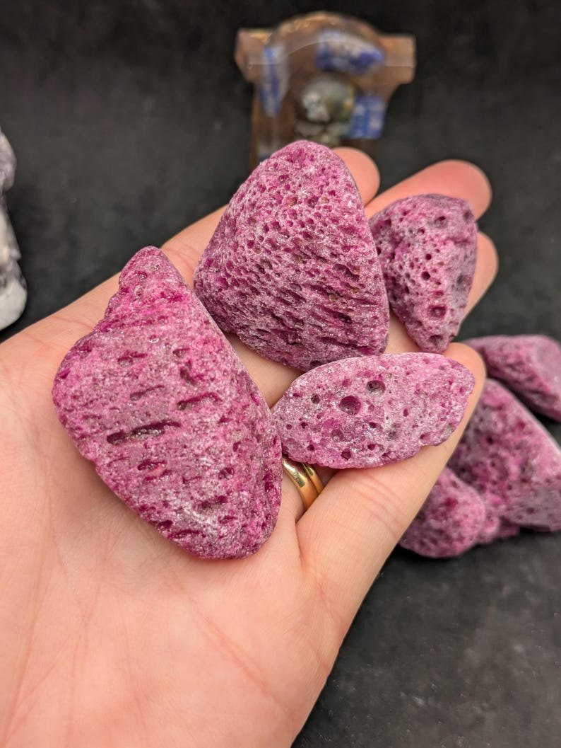 May include: A hand holds several pieces of pink, porous rock. The rocks vary in size and shape, with a textured surface and a deep pink color. The background is dark, highlighting the rocks' color and texture.
