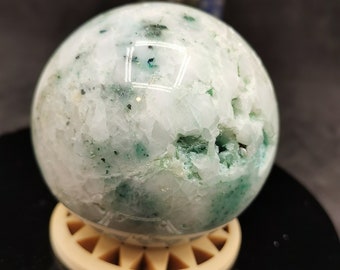 May include: A white and green crystal sphere with a rough, textured surface. The sphere is sitting on a wooden stand.