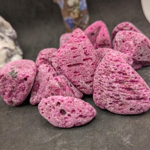 May include: A collection of irregularly shaped, porous, magenta-colored stones. The stones have a rough texture and varying sizes, with some showing small holes. The arrangement is clustered together, creating a natural, organic aesthetic.