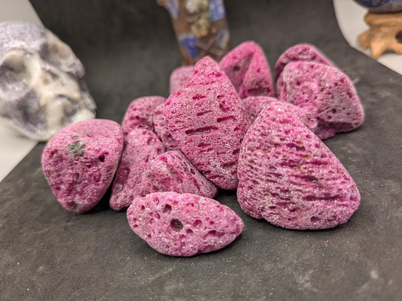 May include: A collection of irregularly shaped, porous, magenta-colored stones. The stones have a rough texture and varying sizes, with some showing small holes. The arrangement is clustered together, creating a natural, organic aesthetic.