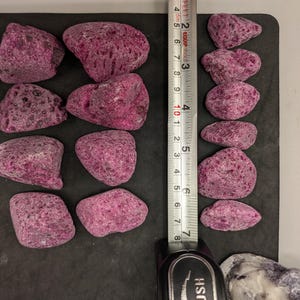 May include: A collection of irregularly shaped, porous, magenta-colored rocks. A ruler shows the rocks are approximately 2 to 3 inches in length. The rocks are arranged on a dark surface, suggesting a decorative or landscaping theme.