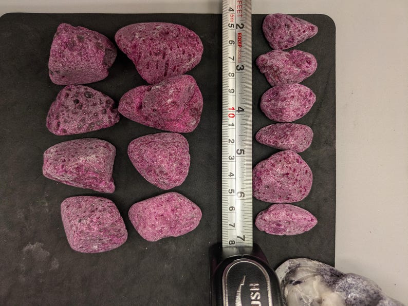 May include: A collection of irregularly shaped, porous, magenta-colored rocks. A ruler shows the rocks are approximately 2 to 3 inches in length. The rocks are arranged on a dark surface, suggesting a decorative or landscaping theme.