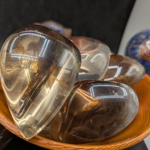May include: A collection of polished, translucent smoky quartz stones in various sizes, resting in a small, round, wooden bowl. The stones have a warm, brown hue and reflect light. A blue and gold decorative sphere is in the background.
