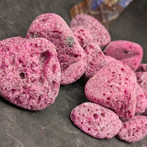 May include: A cluster of irregularly shaped, porous, pink rocks. The rocks have a rough, textured surface with numerous small holes. The color is a vibrant, saturated pink, and they appear to be natural formations.