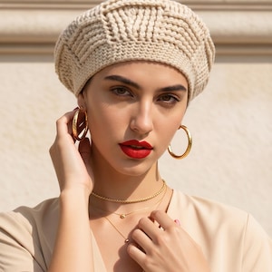 May include: A beige knit beret with a textured pattern. The model is wearing gold hoop earrings and a layered gold necklace. The model has red lipstick and is wearing a beige top.