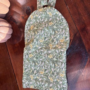 Op de afbeelding: Een stoffen wijnfleszak met een groen en wit bloemenpatroon. De tas heeft een handvat en een herhalend ontwerp van bladeren en gele bloemen. De tas is gemaakt van stof.
