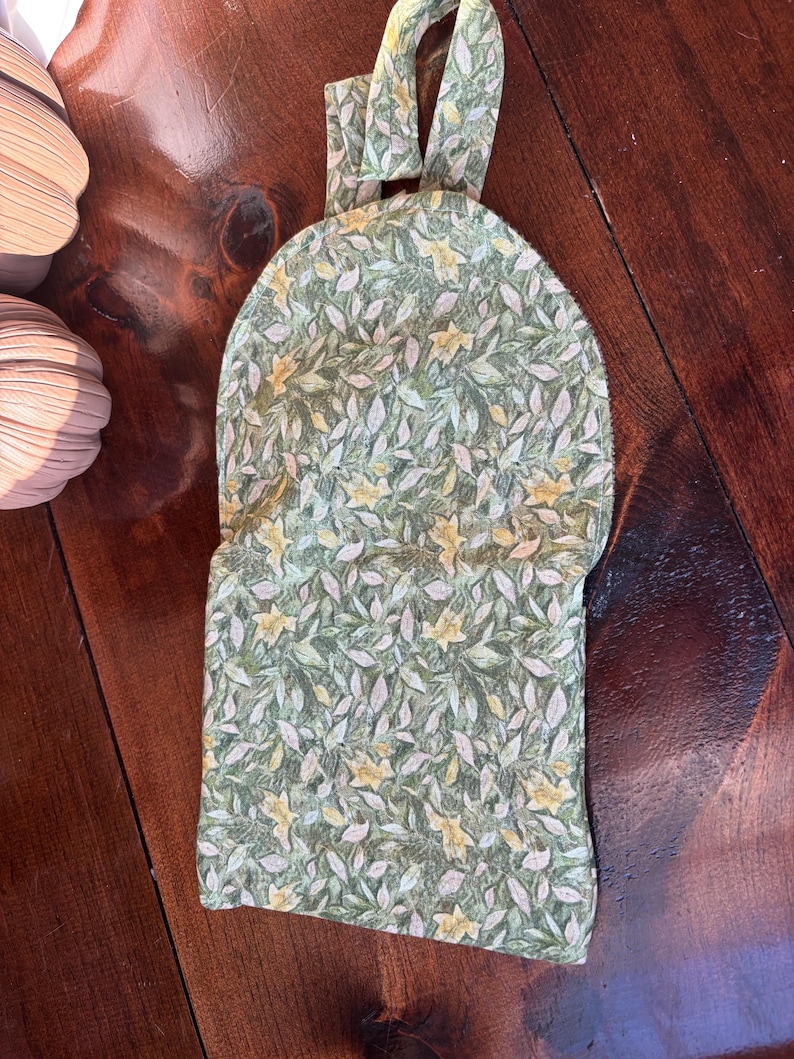 Op de afbeelding: Een stoffen wijnfleszak met een groen en wit bloemenpatroon. De tas heeft een handvat en een herhalend ontwerp van bladeren en gele bloemen. De tas is gemaakt van stof.