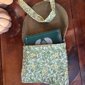 Op de afbeelding: Een kleine, open, groene corduroy tas met een bloemenprint zak en riem. De zak heeft een patroon van groene bladeren en gele bloemen. Een kaartspel is zichtbaar in de tas.