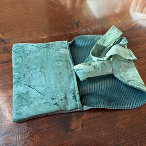 Op de afbeelding: Een teal-kleurige stoffen etui met een gemarmerd patroon op de buitenklep en een corduroy-achtige textuur aan de binnenkant. De etui is open, waardoor de binnenkant zichtbaar is, en heeft een stoffen striksluiting.