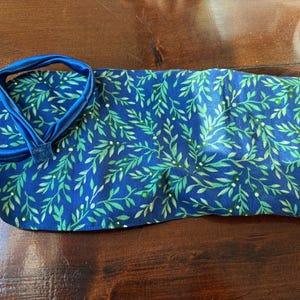 Op de afbeelding: Een blauw stoffen item met een groen bladerpatroon en een blauwe band. De stof is diepblauw met een herhalend patroon van groene bladeren en kleine witte stippen. De band is effen blauw.