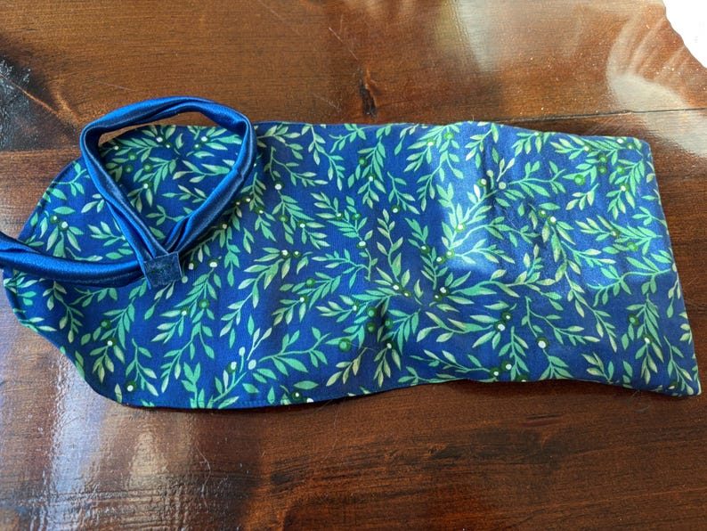Op de afbeelding: Een blauw stoffen item met een groen bladerpatroon en een blauwe band. De stof is diepblauw met een herhalend patroon van groene bladeren en kleine witte stippen. De band is effen blauw.
