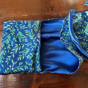 Op de afbeelding: Een blauw satijnen slaapmasker met bijpassende etui. Het masker en de etui hebben een patroon van groene bladeren en witte bessen op een marineblauwe achtergrond. Het masker heeft een blauwe satijnen band.