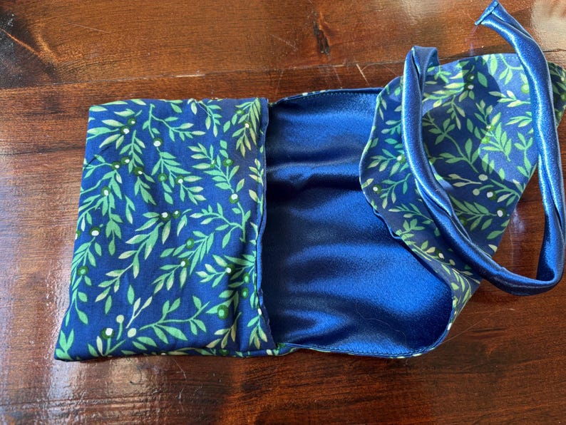 Op de afbeelding: Een blauw satijnen slaapmasker met bijpassende etui. Het masker en de etui hebben een patroon van groene bladeren en witte bessen op een marineblauwe achtergrond. Het masker heeft een blauwe satijnen band.