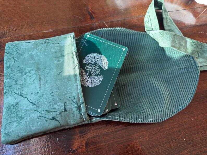 Op de afbeelding: Een groene tarotkaartenset met een wit Keltisch knoopontwerp is gedeeltelijk zichtbaar in een bijpassend groene corduroy etui. De etui is open en onthult de kaarten en een groene kaarthouder van gemarmerde stof.